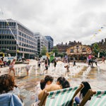 Children play in a city centre fountain, with adults sat around the perimeter in deck chairs.
