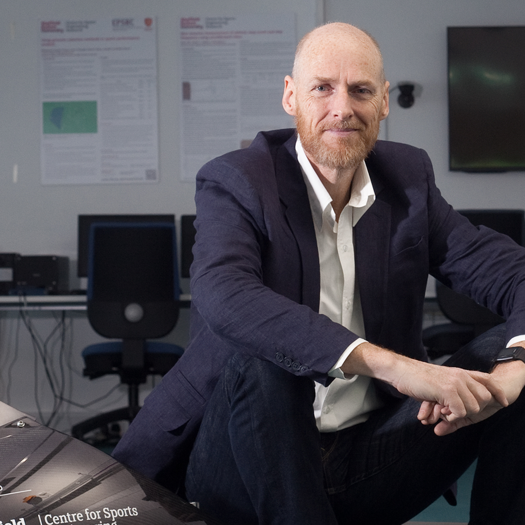 A person wearing a dark navy suit with a white dress shirt, seated on a red cushioned examination table in a clinical setting. A stethoscope hangs around the neck, suggesting a medical professional. The background includes a light blue frosted glass panel and a section of white drawers on the right side.