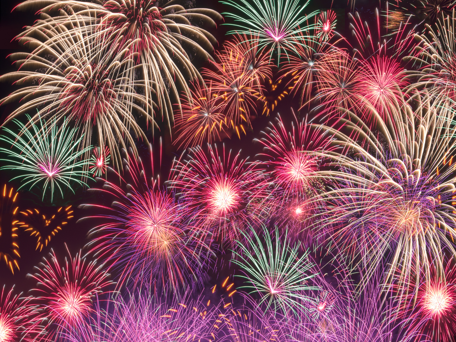 A vibrant display of fireworks bursting against a dark night sky. Multiple colorful explosions in shades of red, pink, purple, green, and gold fill the scene, creating overlapping patterns of light and streaks. The fireworks vary in size and shape, with some forming large circular bursts and others radiating fine trails, giving a festive and celebratory atmosphere.