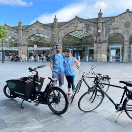 Sheffield Cycle Tours - Two people wearing blue safety vests and helmets standing with bicycles in front of a historic stone building with arched glass entrances. The building has signage for WHSmith, and the foreground shows a cargo bike and two standard bicycles on a paved plaza under a partly cloudy sky.
