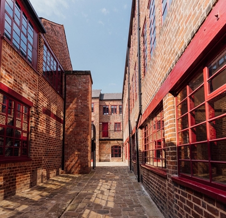 A shot of Leah's Yard, in the sunshine, in the centre of Sheffield. A cobbled path is lined on both sides by renovated red brick industrial buildings.