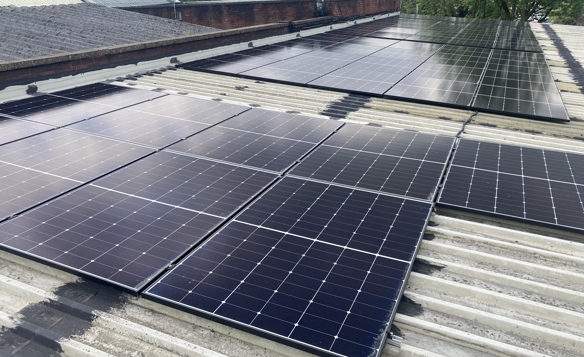 Rows of black solar panels installed on a corrugated metal rooftop, with additional panels visible further back. Surrounding elements include a brick wall, part of a sloped roof, and green trees in the background under a partly cloudy sky.