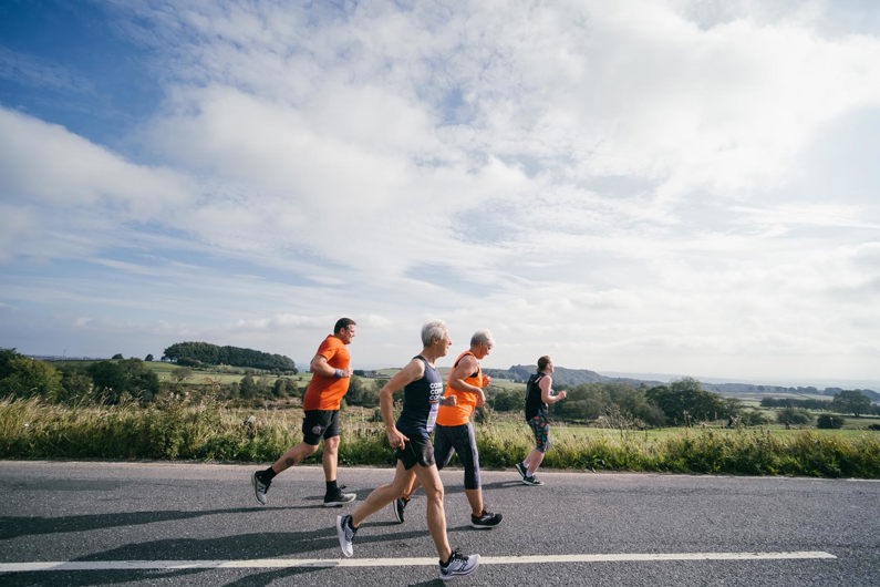 A group of four adults jog along a rural road on a sunny day, with open fields, trees and rolling hills in the background. Two wear bright orange tops and two wear dark athletic clothing. The sky is partly cloudy, and the scene captures a relaxed outdoor run in the countryside.