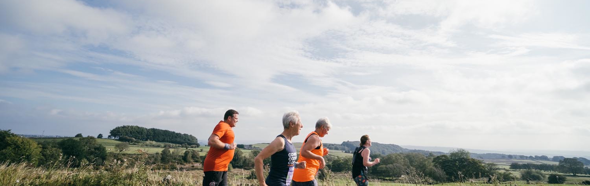 Four people jogging along a rural road on a bright day, with open countryside and green fields stretching into the distance under a partly cloudy sky.