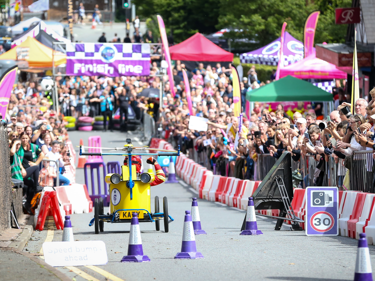 A bright yellow soapbox cart designed like a helicopter races down a city street toward a purple “Finish line” banner. The cart has a rotor on top and blue accents, with the name “KAEY” on the front. Purple-and-white cones mark the track, and red-and-white barriers separate the cheering crowd from the course. Colourful tents and event signage are visible in the background.