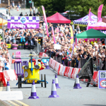 A bright yellow soapbox cart designed like a helicopter races down a city street toward a purple “Finish line” banner. The cart has a rotor on top and blue accents, with the name “KAEY” on the front. Purple-and-white cones mark the track, and red-and-white barriers separate the cheering crowd from the course. Colourful tents and event signage are visible in the background.