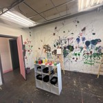 A paint-splattered art room with easels, colourful paint bottles and brushes, and walls covered in expressive splashes, doodles and handprints.