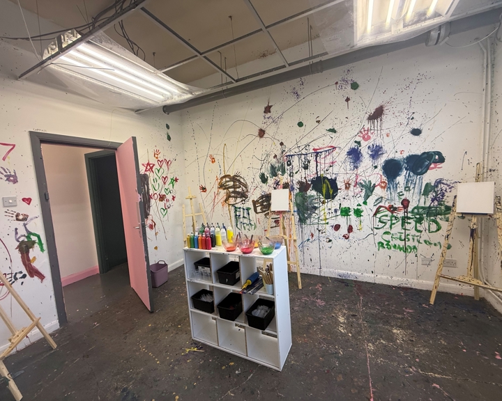 A paint-splattered art room with easels, colourful paint bottles and brushes, and walls covered in expressive splashes, doodles and handprints.
