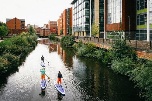 Three people paddleboarding on a narrow river in an urban setting, surrounded by greenery. Modern glass buildings line one side of the river, while older brick buildings are on the other, blending nature with city architecture.