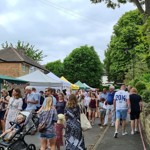 A crowd of people walking along the Nether Edge Farmers' Market. There are stalls situated in front of houses to the left, while the people walk alongside it to the right.