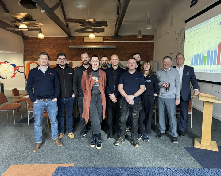 A group of people standing together in a modern meeting room with exposed beams and brick walls. There are orange chairs arranged around the room and a wooden podium on the right. A large screen displays a colourful bar chart with multiple data points. The setting appears to be a business or training event.
