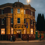 Rutland Arms exterior. The building is red and mustard brick, with the sun setting in the background. The pub is lit up through the windows, with a streetlight lit up at the entrance.