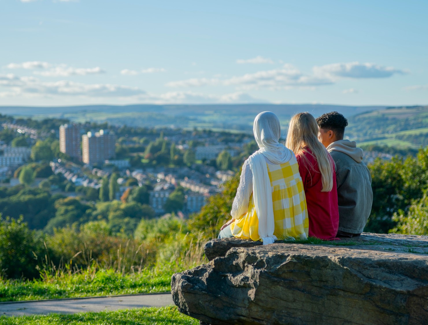 Three people sitting on a stone outcrop overlooking Sheffield, with terraced housing, green hills and open sky stretching across the city landscape.