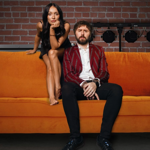 James and Clair Buckley sat on an orange sofa in front of a red brick wall.