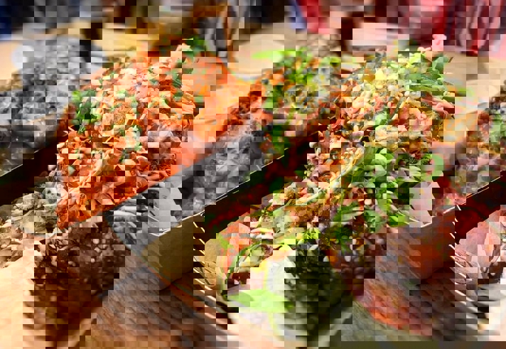 Close-up of two trays of saucy chicken wings served on a wooden table. The tray in the foreground features wings coated in a sticky glaze, topped with sesame seeds, chopped green onions, and a drizzle of creamy sauce, with a lime wedge on the side. The tray in the background contains wings covered in bright orange sauce, garnished with finely chopped herbs. Two small white dipping cups are placed alongside the wings.