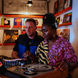 Two people standing at a DJ booth inside a room decorated with vinyl records on shelves. One person is wearing headphones and a vibrant pink patterned shirt with a zebra-print scarf, while the other is adjusting controls on the mixer. A turntable with a colorful slipmat is in the foreground, and framed artwork hangs on the wall above.