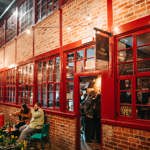 The red brick exterior of Hop Hideout at Leah's Yard, with red painted windows and doors and a black hanging sign that says 'Hop Hideout'. A man and a woman are sat on a table outside drinking, the door is open and customers are waiting at the bar.
