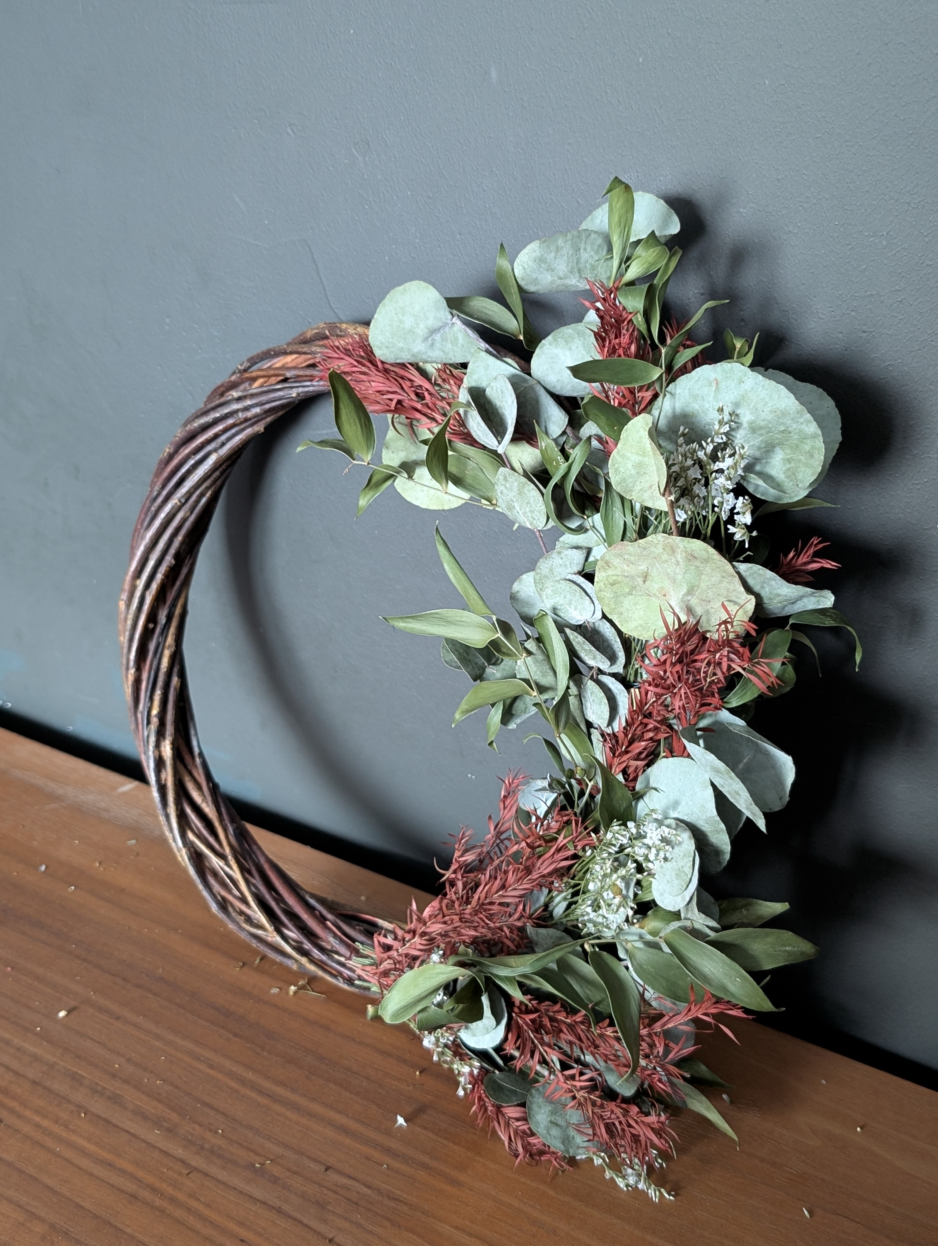A half-covered wreath featuring a twisted vine base adorned with clusters of dried eucalyptus leaves, red foliage, and small white flowers. The arrangement is displayed on a wooden surface against a dark grey wall, showcasing an organic and asymmetrical design.