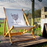 A large wooden deckchair printed with the Old Horns Inn logo stands on a raised platform overlooking rolling green countryside, with a historic stone church visible in the background under a cloudy sky.