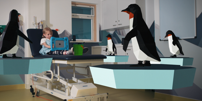 A child is lying in a bed in a hospital ward. There are 3D models of penguins around the room. The child is entertaining herself with a tablet type device.
