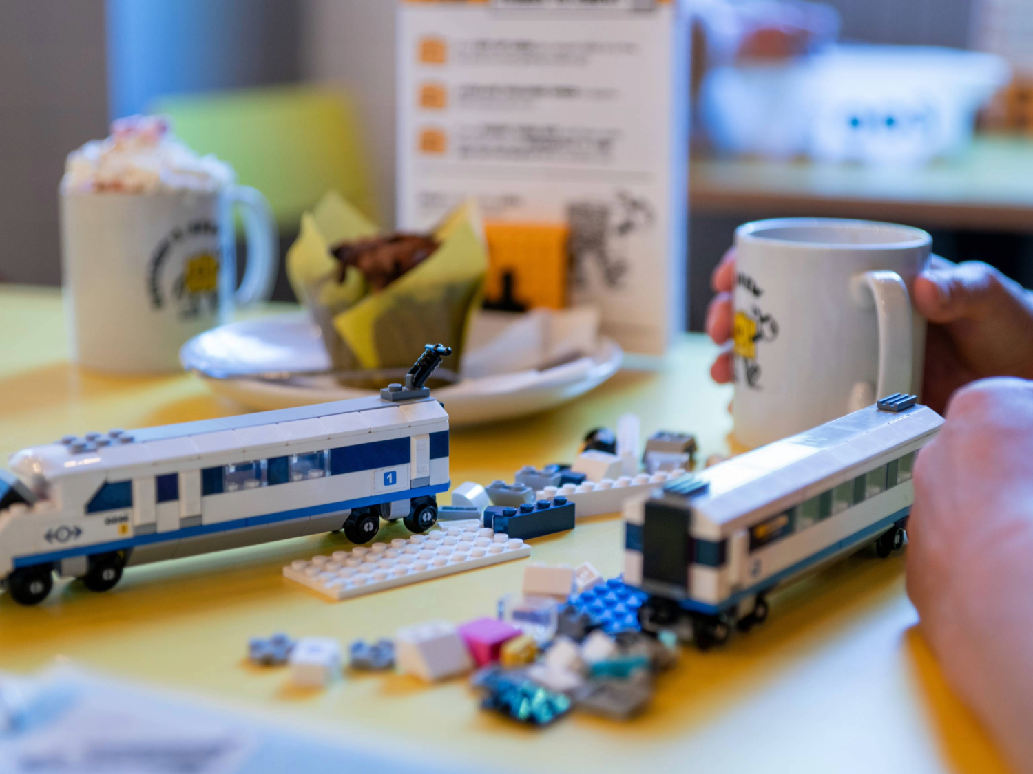 A wooden table in a cafe. On the table are two hot drinks and a partially built Lego© train, along with the instructions and the remaining pieces.