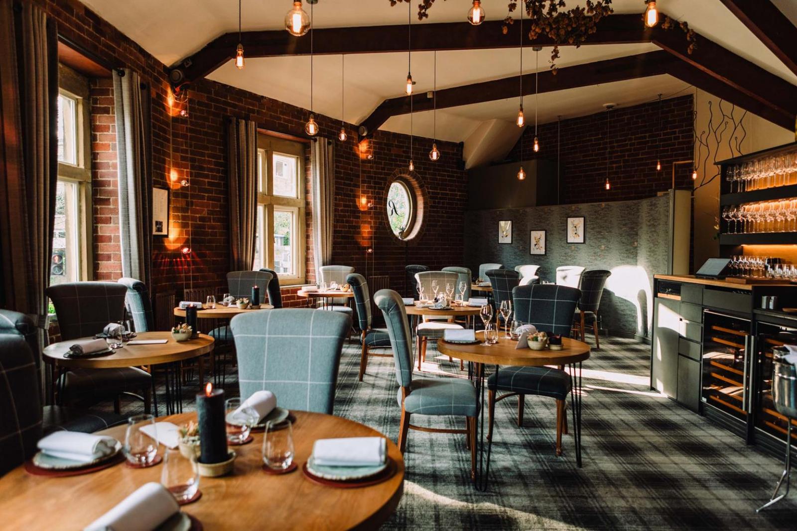 An elegant restaurant interior featuring round wooden tables set with folded napkins, glassware, and small decorative items. The space has exposed brick walls, large windows with gray curtains, and a patterned carpet. Pendant lights hang from the ceiling beams, creating warm ambient lighting. A wine storage area and shelving with glasses are visible on the right side of the room, adding to the sophisticated atmosphere.