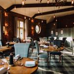 An elegant restaurant interior featuring round wooden tables set with folded napkins, glassware, and small decorative items. The space has exposed brick walls, large windows with gray curtains, and a patterned carpet. Pendant lights hang from the ceiling beams, creating warm ambient lighting. A wine storage area and shelving with glasses are visible on the right side of the room, adding to the sophisticated atmosphere.