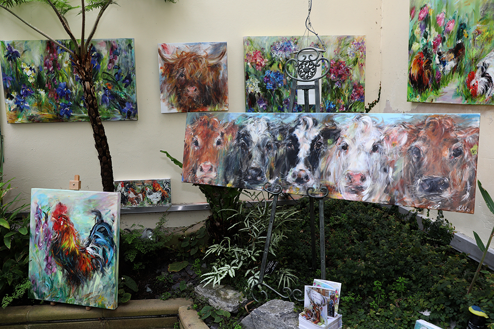 A colourful art display featuring several vibrant animal paintings arranged against a light wall and surrounded by greenery. The largest canvas on an easel shows close-up portraits of cows in expressive brushstrokes. Other paintings depict a rooster and more cows among bright flowers. Small prints and cards are placed on a stone ledge in front, creating a lively and nature-inspired setting.