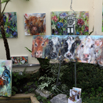 A colourful art display featuring several vibrant animal paintings arranged against a light wall and surrounded by greenery. The largest canvas on an easel shows close-up portraits of cows in expressive brushstrokes. Other paintings depict a rooster and more cows among bright flowers. Small prints and cards are placed on a stone ledge in front, creating a lively and nature-inspired setting.