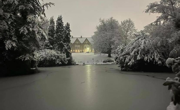 Snow-covered landscape with a large frozen pond in the foreground, surrounded by trees heavily laden with snow. In the background, a stately building with warm lights glowing from its windows stands at the centre, partially framed by the snowy trees. The sky is overcast, creating a serene winter atmosphere.