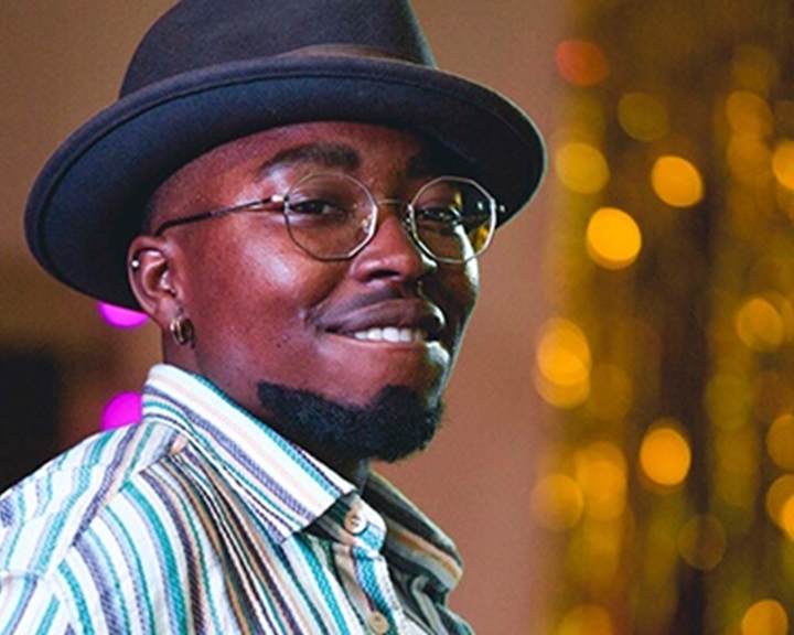 A man of African heritage, wearing a pork pie hat and a striped shirt, smiles broadly.