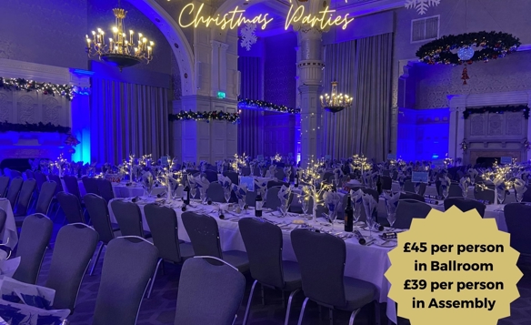 Elegant ballroom set up for a Winter Wonderland Christmas party, featuring long banquet tables with white tablecloths, festive centerpieces with twinkling lights, and neatly arranged chairs. The room is decorated with garlands, wreaths, and chandeliers, illuminated by blue accent lighting. Text on the image reads “Winter Wonderland Christmas Parties” and a gold badge displays pricing: £45 per person in Ballroom, £39 per person in Assembly. Crowne Plaza logo appears at the bottom.