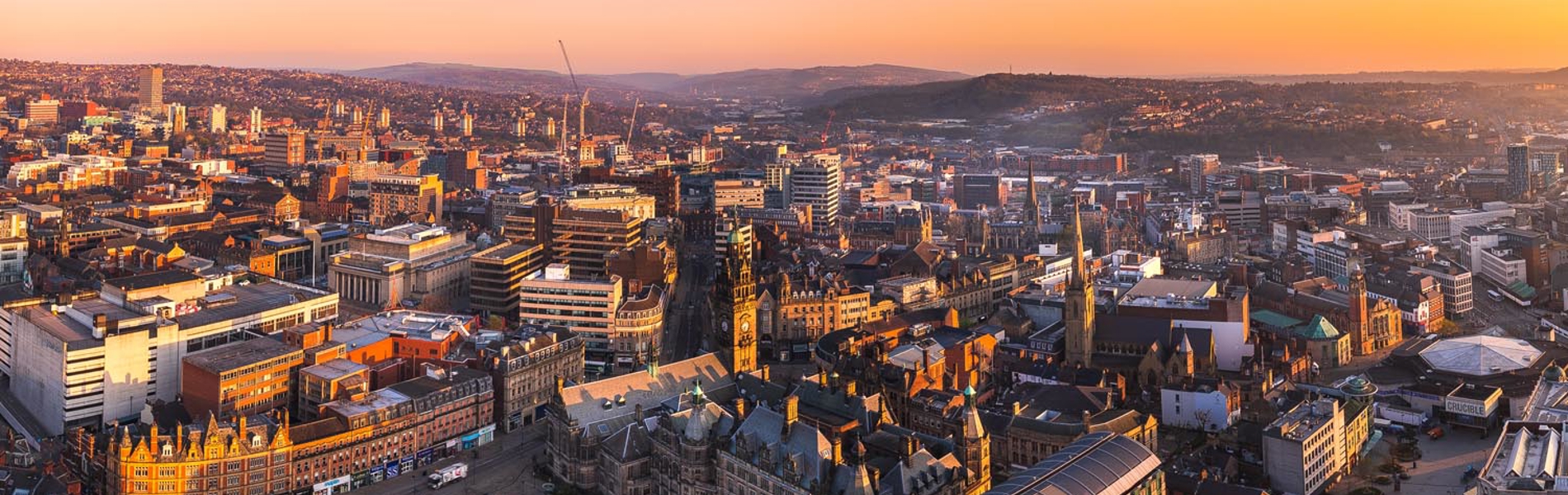 A panoramic view of Sheffield at sunrise, showcasing a mix of historic and modern architecture. The foreground features the ornate Sheffield Town Hall with its clock tower, surrounded by curved streets and green spaces. Beyond, densely packed buildings stretch across the city, with rolling hills visible in the distance under a vibrant orange and purple sky.