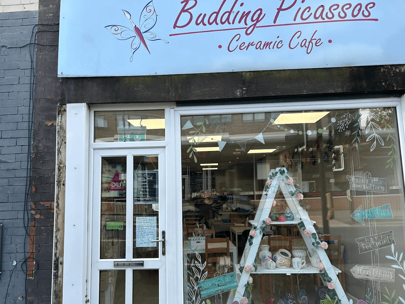 Exterior of Budding Picassos Ceramic Café with a light blue sign, white shopfront, large display window, and decorative painted details.