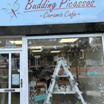 Exterior of Budding Picassos Ceramic Café with a light blue sign, white shopfront, large display window, and decorative painted details.
