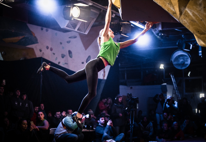 A woman  is on a indoor climbing wall. She is hanging on by a single hand and is reaching for another hand-hold. There is a large crowd  watching.