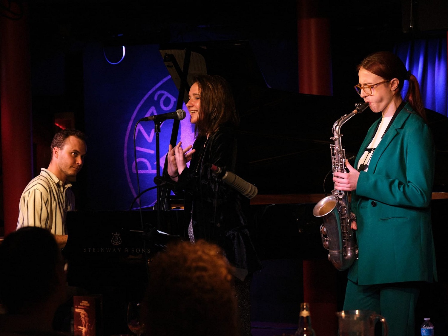 Three people performing music on a stage, one playing piano, one playing saxophone and one singing.