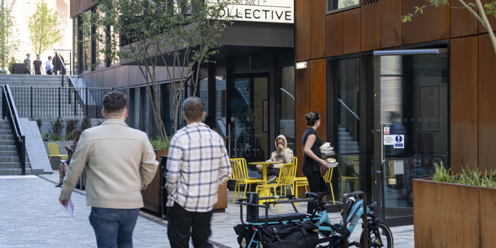Two people walking along a paved pedestrian street lined with modern buildings featuring large windows and rust-colored panels. In the foreground, a cargo-style electric bike is parked near a planter. To the right, a café with bright yellow chairs and tables is visible, with one person seated and another carrying drinks. A sign above the café entrance reads “COLLECTIVE.” Trees and steps leading to an upper level appear in the background.