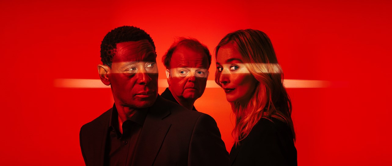 Three people stand closely together against a vivid red background, lit by a horizontal streak of light that cuts across the scene. They wear dark clothing, creating a strong contrast with the bright, atmospheric backdrop.