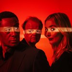 Three people stand closely together against a vivid red background, lit by a horizontal streak of light that cuts across the scene. They wear dark clothing, creating a strong contrast with the bright, atmospheric backdrop.