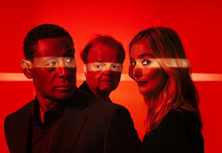 Three people stand closely together against a vivid red background, lit by a horizontal streak of light that cuts across the scene. They wear dark clothing, creating a strong contrast with the bright, atmospheric backdrop.