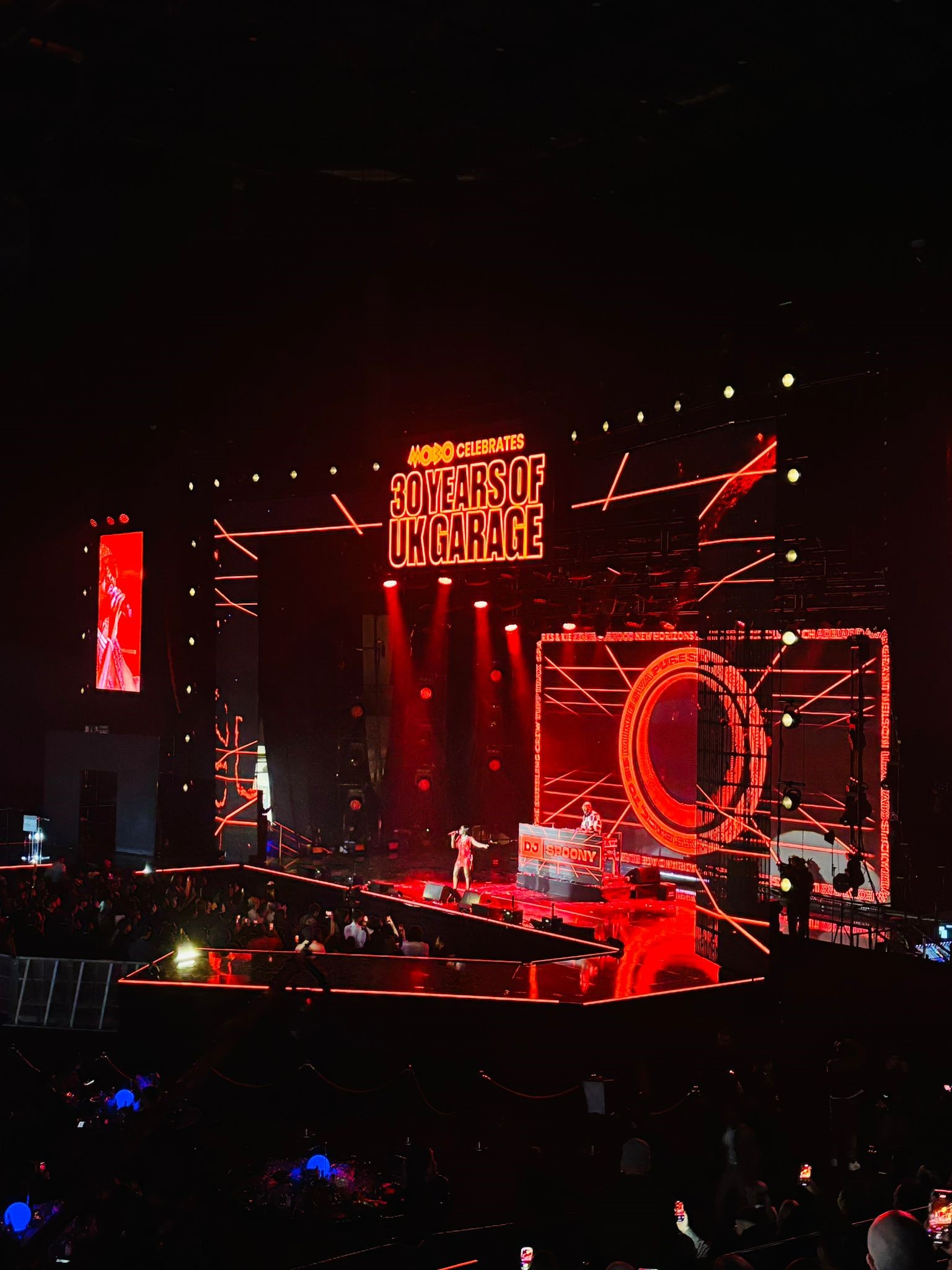 A large concert stage illuminated in red lights with geometric patterns and LED screens. At the top, bold text reads “MOBO Celebrates 30 Years of UK Garage.” A performer stands at the front of the stage, and a DJ booth labeled “DJ Spoony” is positioned behind. The audience is visible in the foreground, with spotlights and glowing wristbands creating a vibrant atmosphere inside a dark arena.