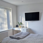 A modern bedroom featuring light linen, a wall-mounted flat-screen TV, a desk with a window.