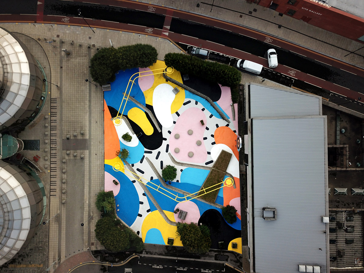 Aerial view of a colorful urban courtyard featuring an abstract geometric mural on the ground with bold shapes in yellow, blue, pink, orange, black, and white. The space includes scattered trees, circular seating areas, and is surrounded by modern buildings and a curved road with parked vehicles.