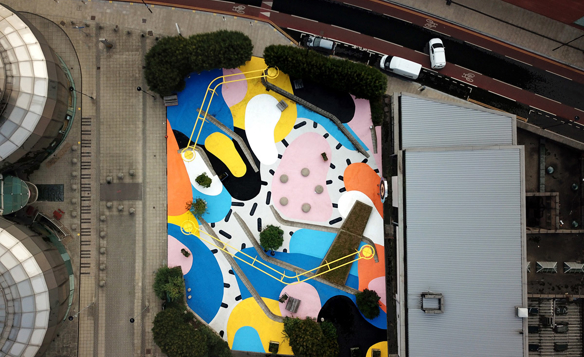 Aerial view of a colorful urban courtyard featuring an abstract geometric mural on the ground with bold shapes in yellow, blue, pink, orange, black, and white. The space includes scattered trees, circular seating areas, and is surrounded by modern buildings and a curved road with parked vehicles.