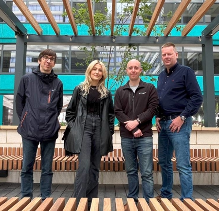 Four people standing together in an outdoor courtyard area with modern architecture. The setting features wooden benches arranged in rows, a pergola-style structure overhead, and a small tree planted in the center. Behind them is a multi-story building with teal panels and large windows, displaying signage that reads “Pennine Five” and “Offices To Let.”