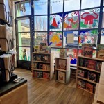 Interior view of Hillsborough Bookshop looking toward the front windows decorated with large, colourful illustrations. Small wooden book displays filled with children’s books stand on the wooden floor, and bunting hangs near the doorway.