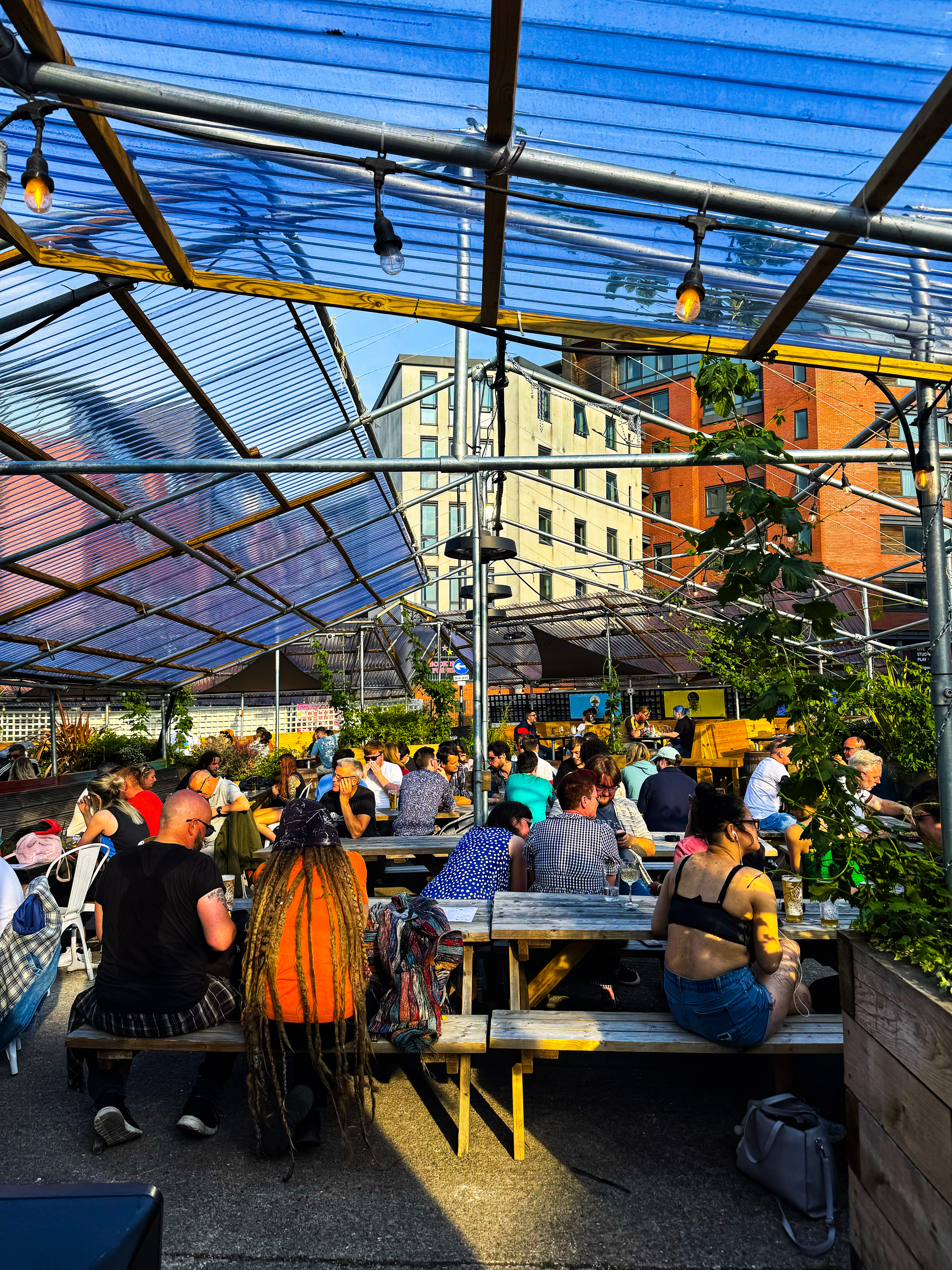  A busy outdoor seating area filled with people at wooden benches under a transparent canopy. The sun shines brightly overhead, casting strong light and highlighting surrounding greenery.