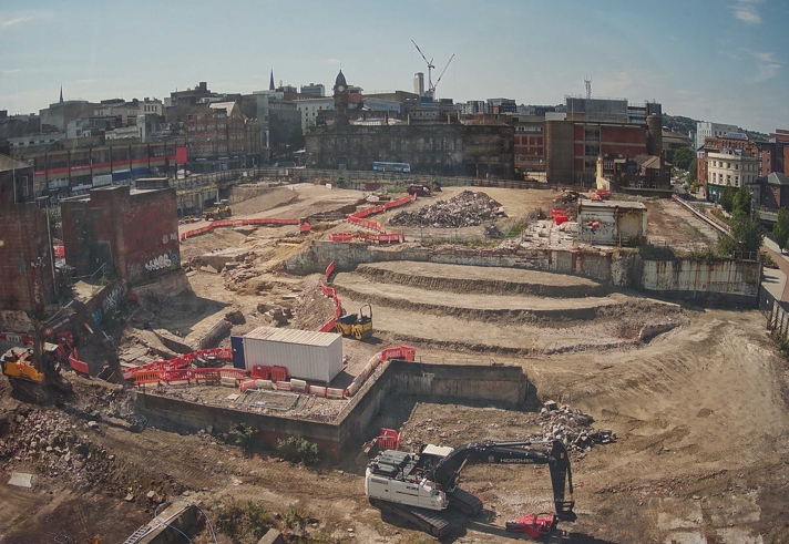 Construction site in an urban area with terraced excavation levels, machinery, and red safety barriers. Surrounding the site are older brick buildings and cranes in the background under a clear blue sky.