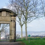 A historic stone archway with decorative carvings stands in a park surrounded by bare trees. The arch frames a distant view of a cityscape with hills in the background. Yellow daffodils bloom in the grassy area near the arch, and a small green sign is visible on the lawn. The sky is bright with scattered clouds, creating a clear and open atmosphere.